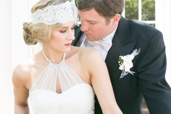 A bride in a white dress and lace headpiece stands next to a groom in a black tuxedo with a white bow tie and boutonniere.