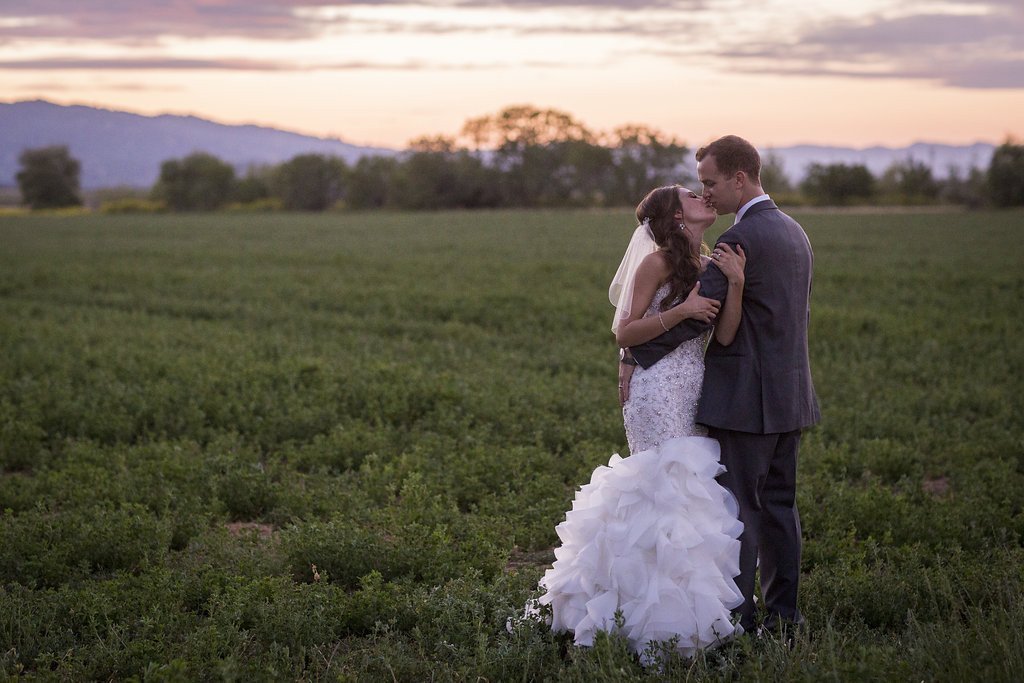 A bride and groom embrace in a field at sunset, with the bride wearing a white gown and the groom in a suit. Trees and mountains are visible in the background.