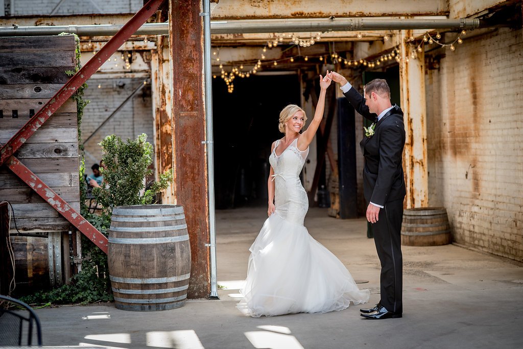 A bride and groom dance in an industrial-style venue with exposed metal beams, barrel decorations, and string lights.