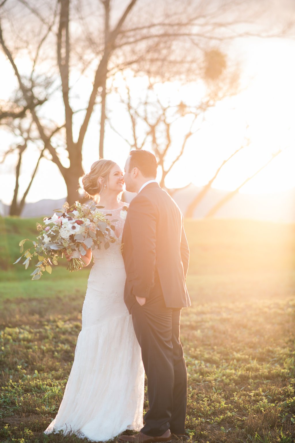 A bride and groom stand closely in a sunlit field, with the bride holding a bouquet and the groom wearing a suit.