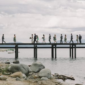 A wedding party walks in a line across a wooden pier over a calm body of water under a cloudy sky.