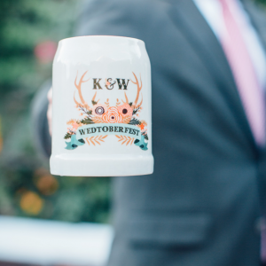 A person holds a white mug with a floral design and antlers, labeled "K&W WEDTOBERFEST." Their face and the background are blurred, focusing on the mug.