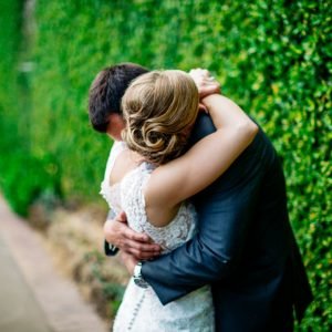 A couple embraces in front of a green hedge, with the woman wearing a white lace dress and the man in a suit.