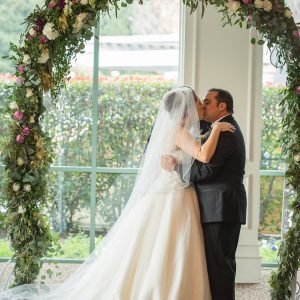 A bride in a white gown and veil kisses a groom in a black suit under a floral arch during their wedding ceremony.