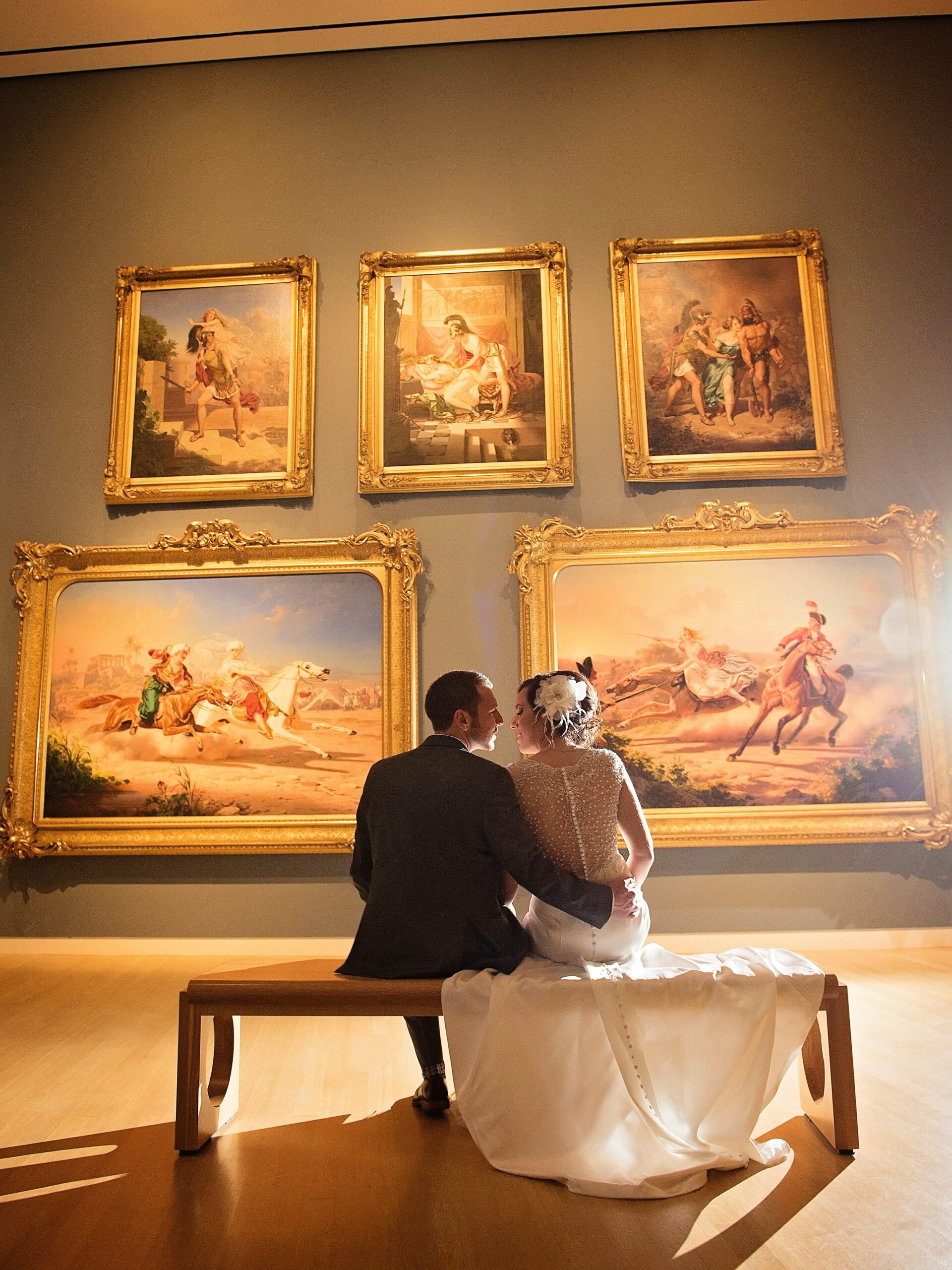 A bride and groom sit on a bench in an art gallery, facing away from the camera and looking at a wall displaying six large, framed paintings.