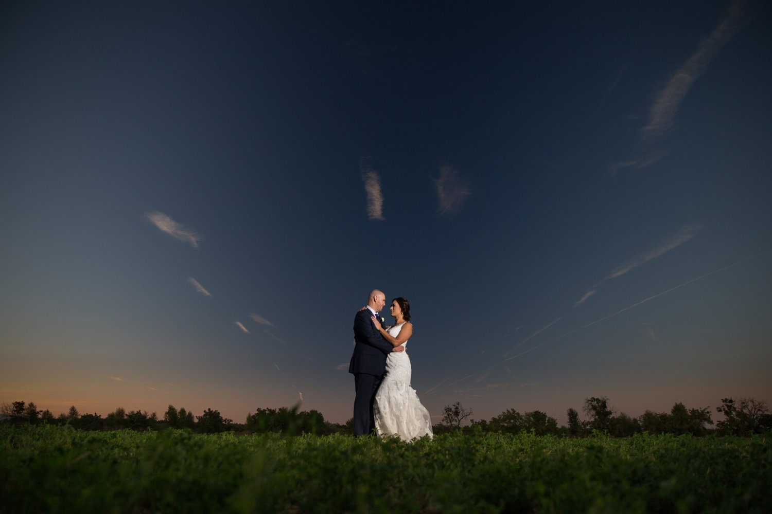 A bride and groom stand outdoors under an evening sky, embracing each other with a vast, dark blue sky and a hint of sunset in the background.