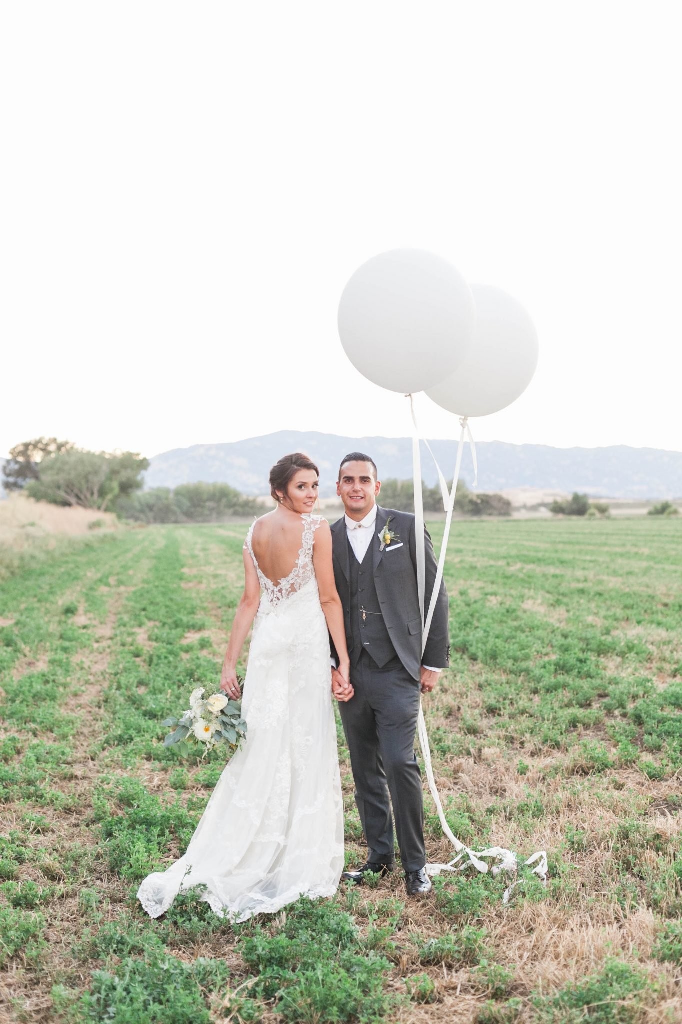 A bride in a white dress and a groom in a gray suit stand side by side holding hands and large white balloons in a grassy field with hills in the background.