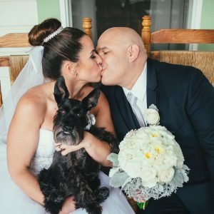 A bride and groom share a kiss while holding a black dog. The bride holds a bouquet of white flowers.