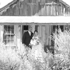 A couple dressed in formal attire stands in front of an old wooden building, surrounded by overgrown plants. The woman holds a bouquet and they both appear ready for a special occasion.