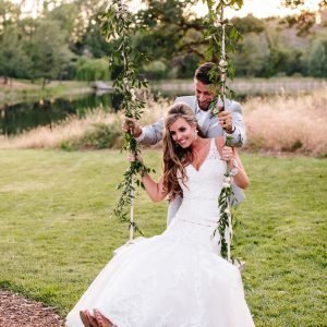 A bride in a white gown sits on a swing decorated with greenery, smiling while holding the ropes. A groom in a light suit stands behind her, both outdoors with a grass and tree-filled background.