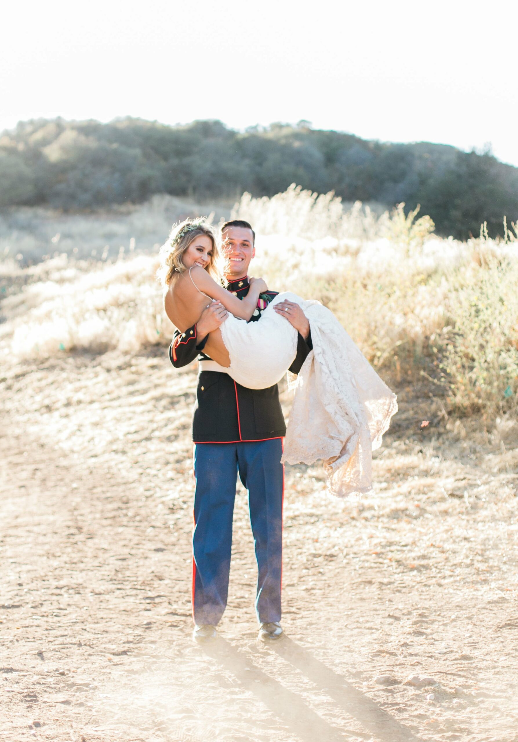 A man in a military uniform carries a woman in a white wedding dress in an open field with hills and dry vegetation in the background.