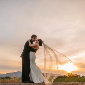 A bride and groom embrace and kiss outdoors during sunset, with the bride's veil flowing in the wind.