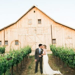 A couple in wedding attire stands close together in a vineyard, positioned in front of a large wooden barn.