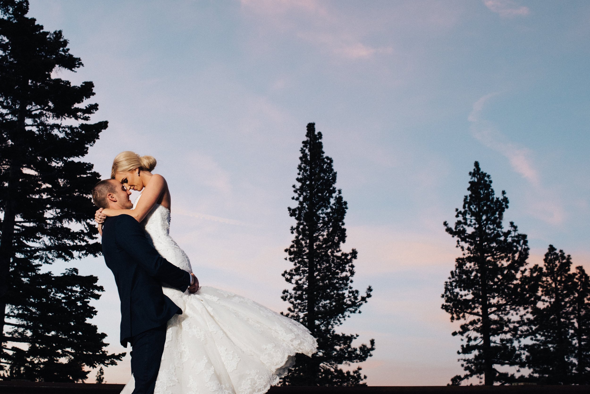 A groom lifts the bride into the air, both smiling, with tall pine trees and a dusk sky in the background.