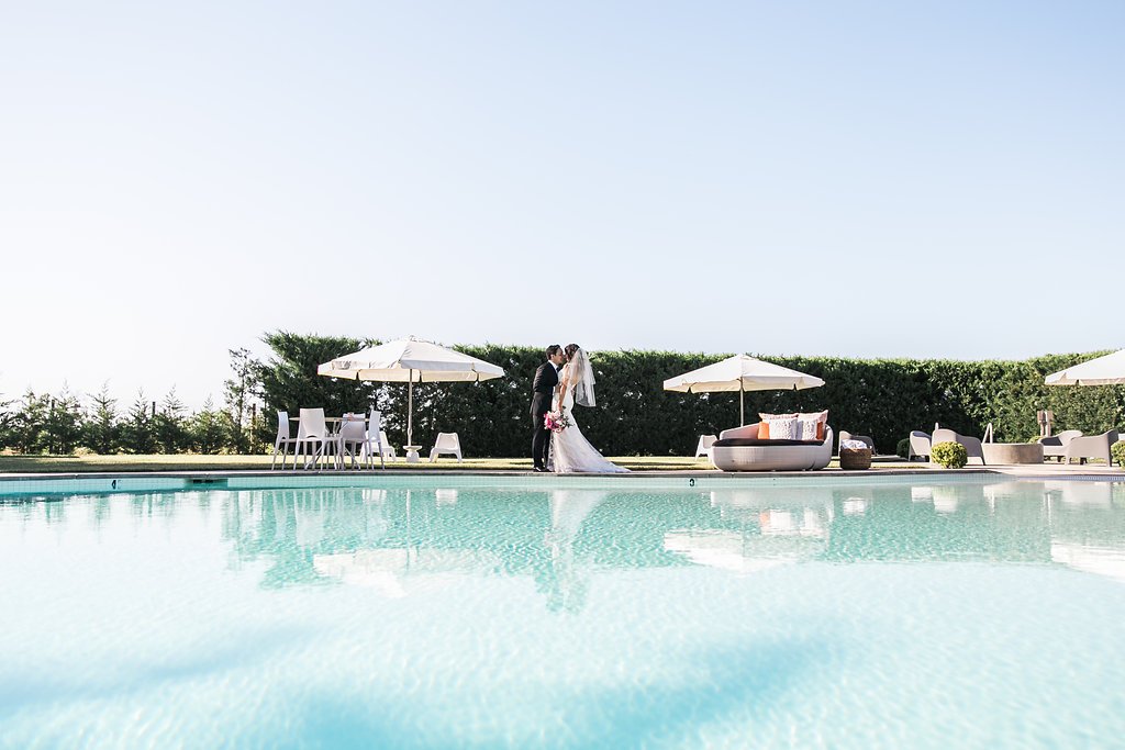 A bride and groom stand facing each other by a poolside, with white umbrellas, chairs, and a hedge in the background.