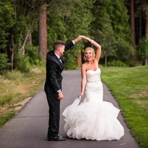 A couple in formal attire dance on a path surrounded by trees and grass, with the bride in a white gown and the groom in a black suit.