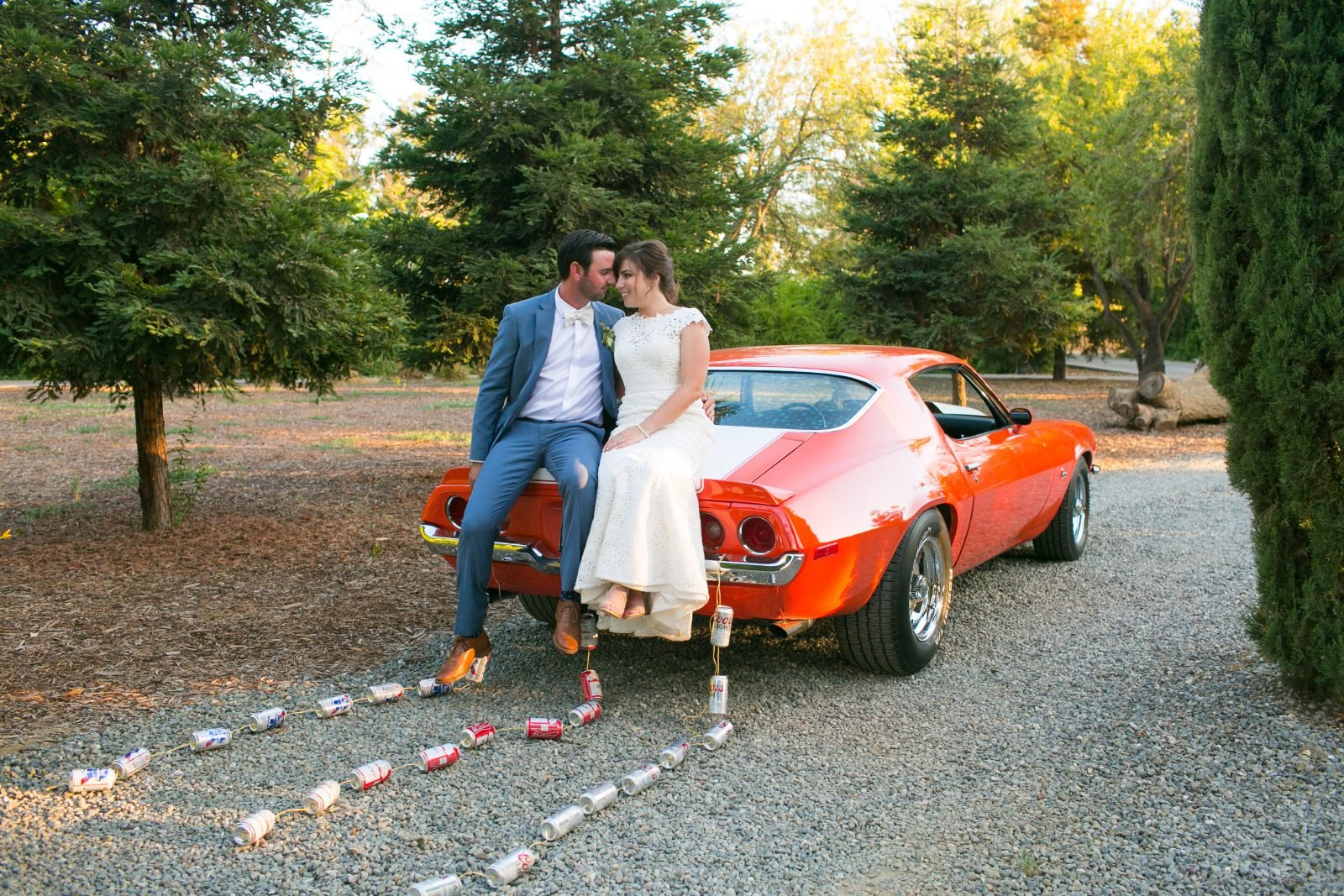 A couple dressed formally sits on the trunk of an orange classic car decorated with cans tied to the back, parked on a gravel path in a wooded area.