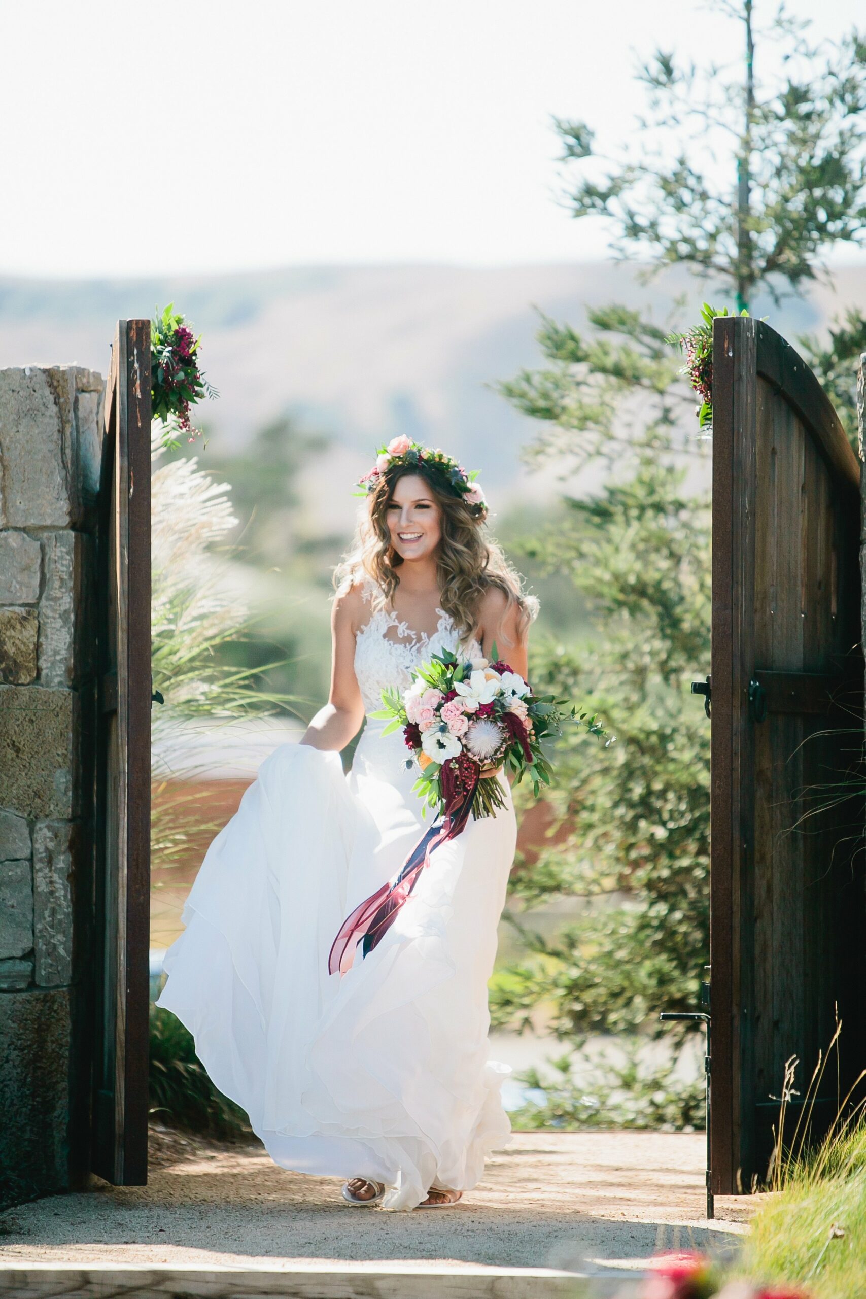 A bride in a white gown and floral crown enters through a wooden gate, holding a colorful bouquet in a sunlit outdoor setting.