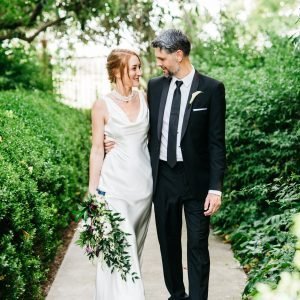 A couple dressed in wedding attire walks arm-in-arm down a garden path. The bride holds a bouquet and wears a white dress, while the groom wears a black suit with a white shirt and tie.