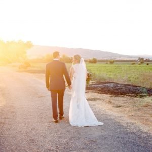 A bride and groom holding hands walk down a sunlit dirt path, surrounded by fields and hills in the background.