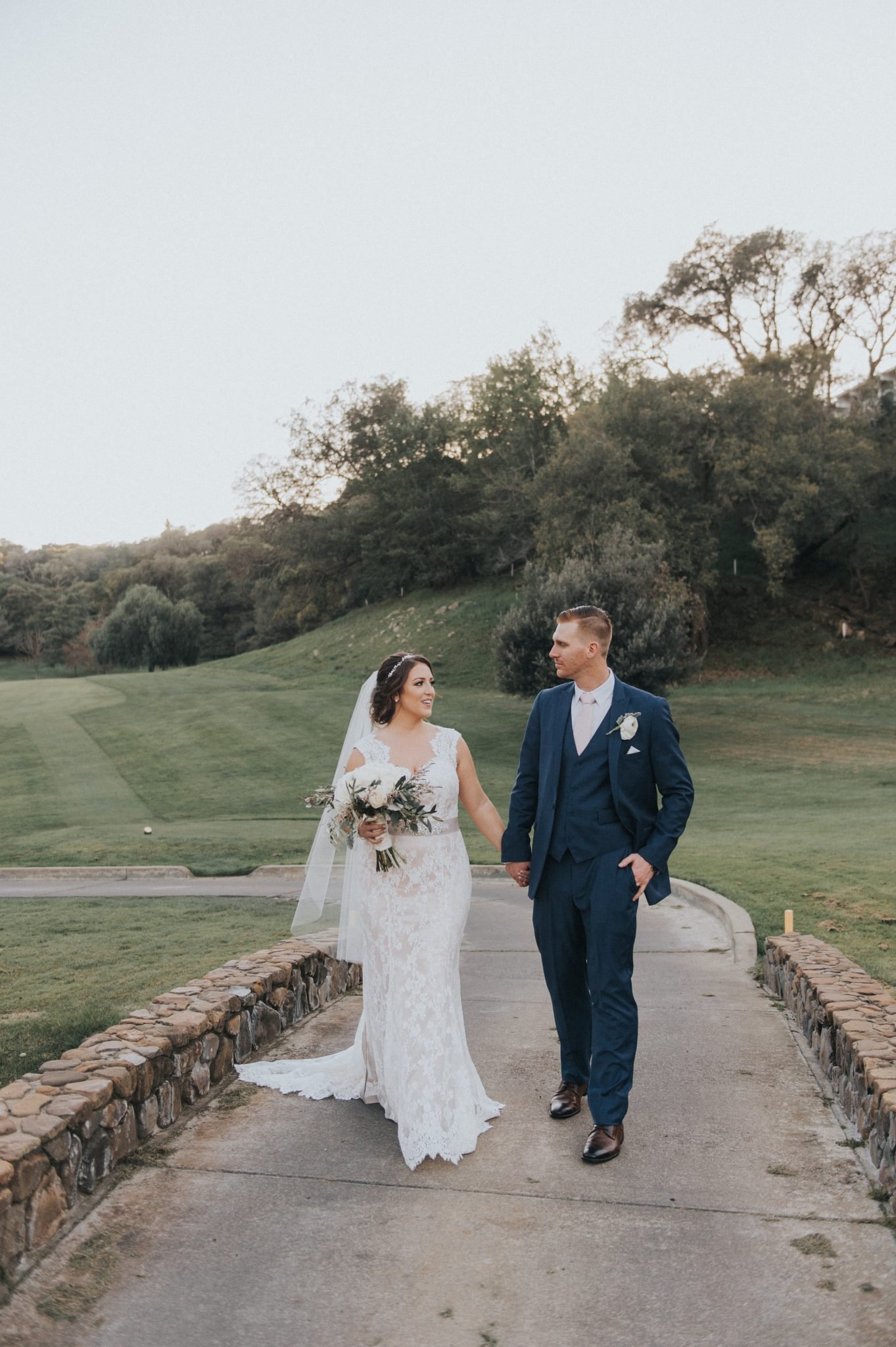 A couple dressed in wedding attire walks hand in hand on a paved path through a lush, green landscape.