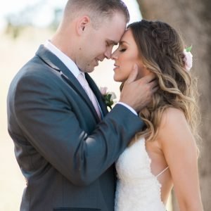 A couple embracing closely with their foreheads touching outdoors, the man wears a gray suit and the woman wears a white dress.