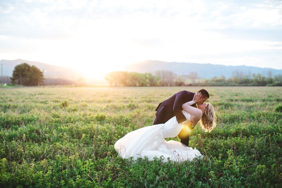 A couple dressed in wedding attire share a kiss in a grassy field during sunset, with mountains visible in the background.