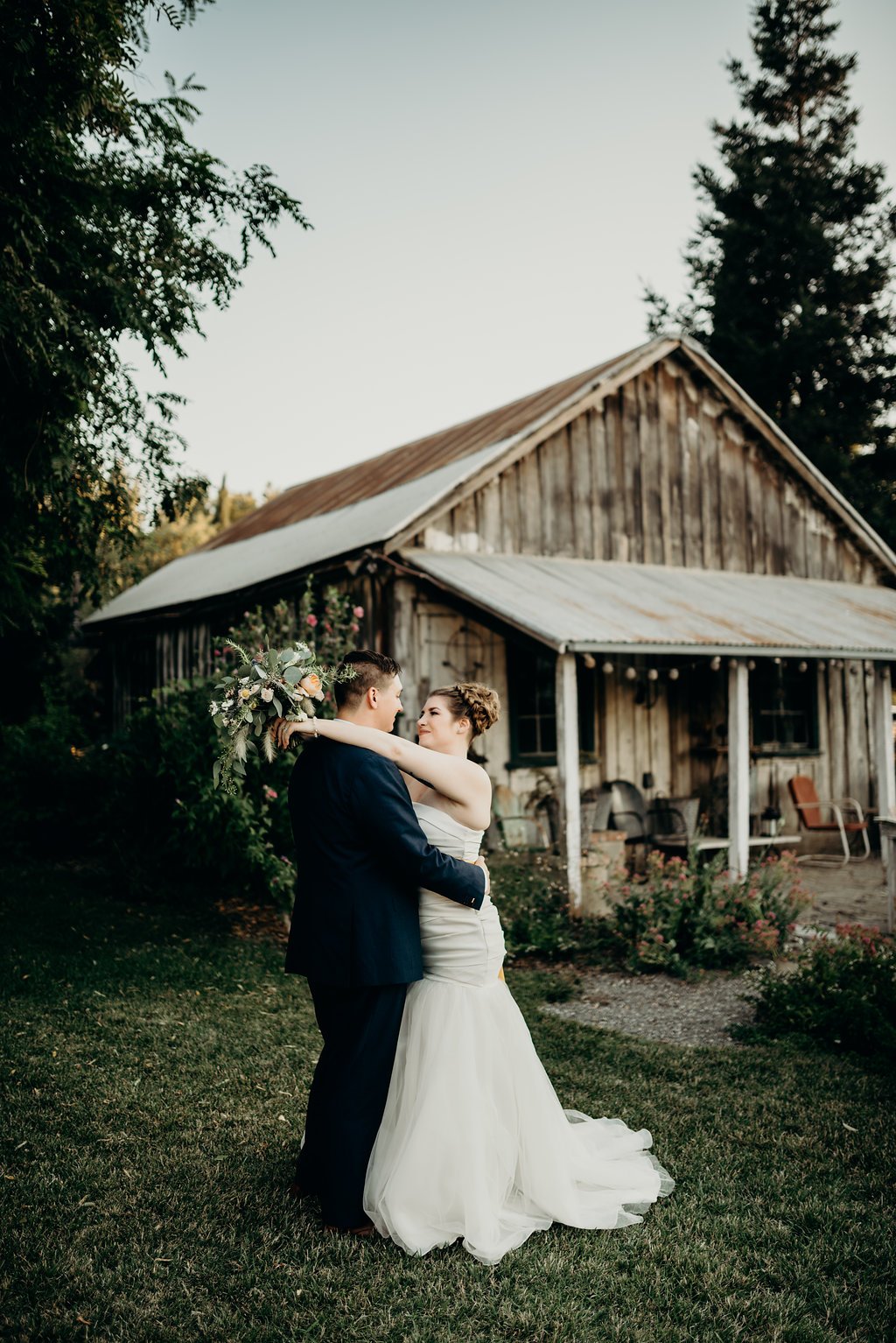 A couple in wedding attire shares an embrace in front of a rustic wooden building with a corrugated metal roof.