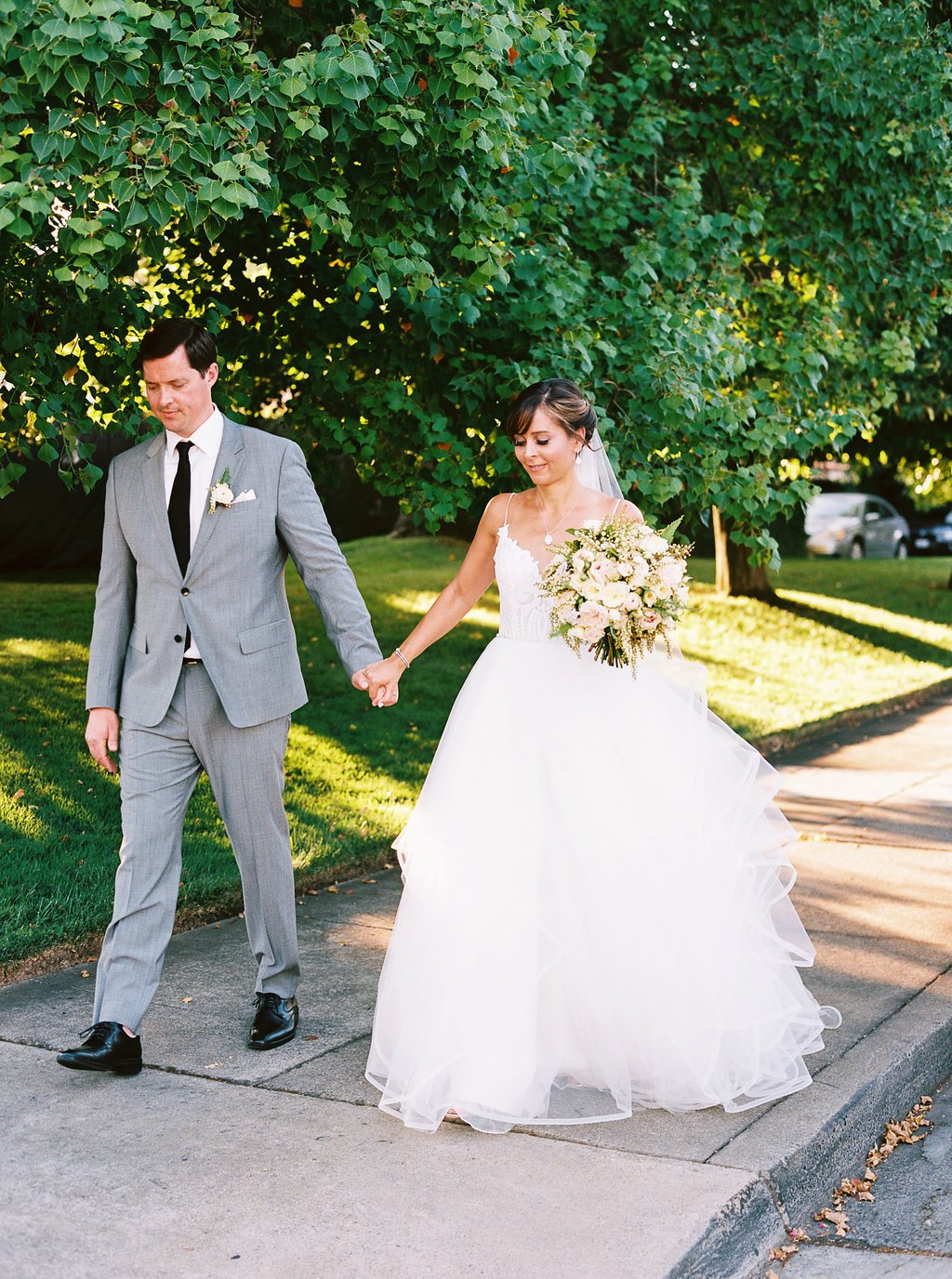 A couple walks hand-in-hand on a sunny sidewalk wearing wedding attire; the groom in a grey suit, the bride in a white gown with a bouquet. Trees and grass in the background.