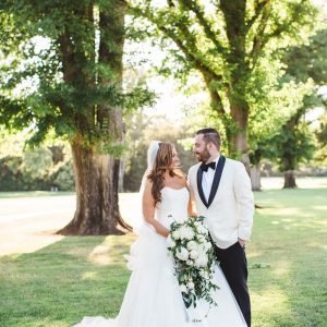 A couple in wedding attire stands outdoors on a sunny day; the bride is holding a bouquet of white flowers, and the groom is wearing a white jacket with black lapels. Trees are visible in the background.