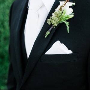 Close-up of a person wearing a black suit, white shirt, and white tie, with a white pocket square and a boutonniere in the jacket's lapel.