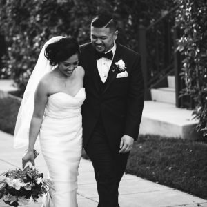 A bride and groom walk together outdoors. The bride holds a bouquet and wears a strapless dress and veil. The groom wears a suit with a bow tie. Both are smiling.