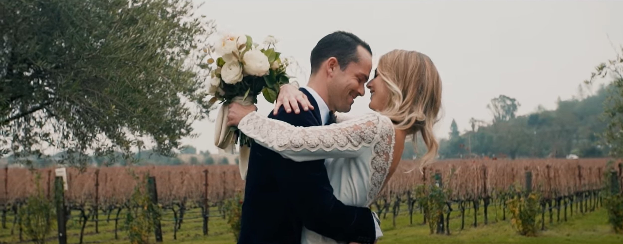 A smiling couple in wedding attire embraces in a vineyard, with the bride holding a bouquet of white flowers. The sky is overcast.