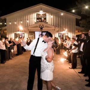 A couple shares a kiss surrounded by guests holding sparklers outside a brightly lit barn during an evening celebration.