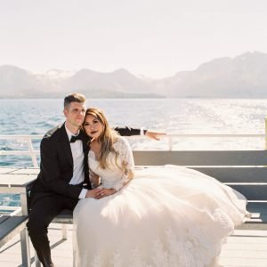 A couple in wedding attire sits on a bench outdoors with a large body of water and mountains in the background. They are holding hands.