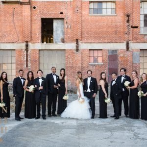 A wedding party poses in formal attire against a brick building backdrop, with the bride and groom at the center surrounded by bridesmaids in black dresses and groomsmen in black tuxedos.