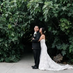 A couple dressed in wedding attire stands in front of dense greenery. The bride is embracing the groom from behind, both smiling. The groom is wearing a black suit; the bride is in a white gown.