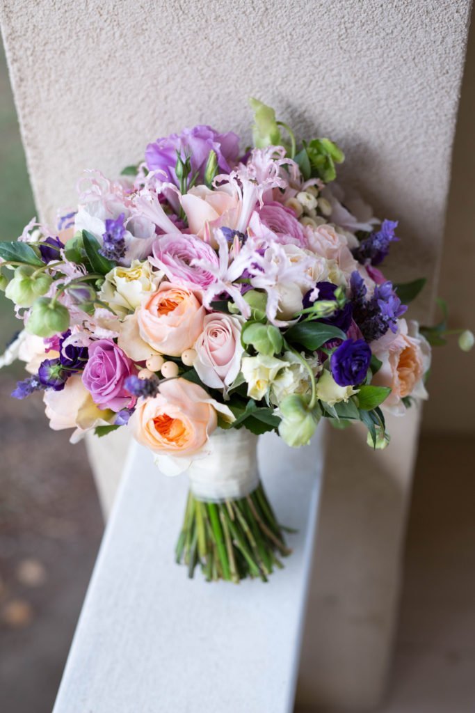 A mixed bouquet of flowers, including pink roses, purple and white blooms, and green leaves, is displayed on a ledge.