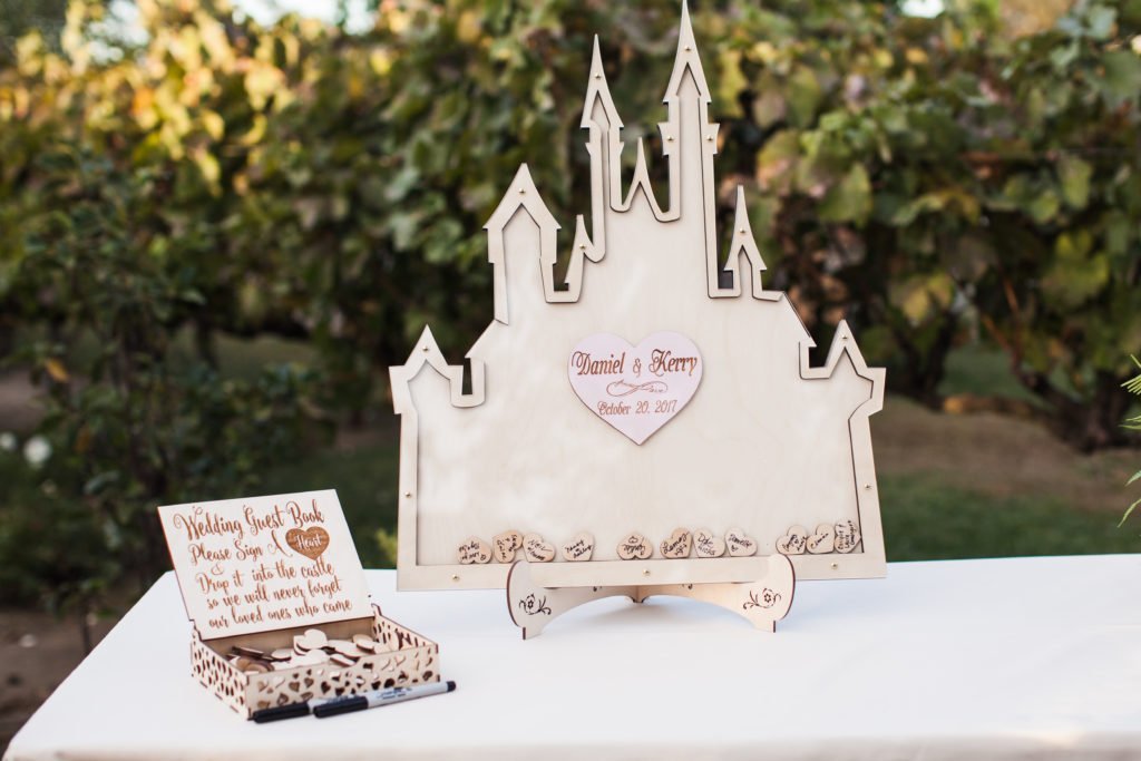 A wooden guest book shaped like a castle with small heart-shaped tokens sits on a table. Another wooden piece instructs guests to sign the tokens for a decorative wedding guest book display.