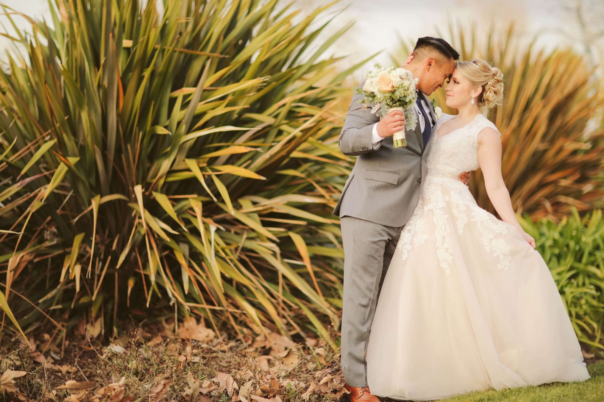 A couple in wedding attire stands close together outdoors. The groom, holding a bouquet, kisses the bride's forehead. They are surrounded by greenery and plants.