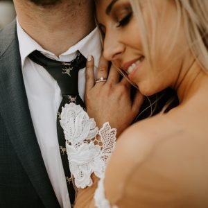 A woman in a wedding dress embraces a man in a suit, showing a close-up of their torsos. The woman's hand, adorned with a ring, gently holds the man's tie.