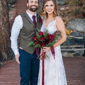 A man in a vest and tie stands next to a woman in a white lace dress holding a bouquet of red flowers. They both smile at the camera, standing on a wooden path with trees and stones in the background.