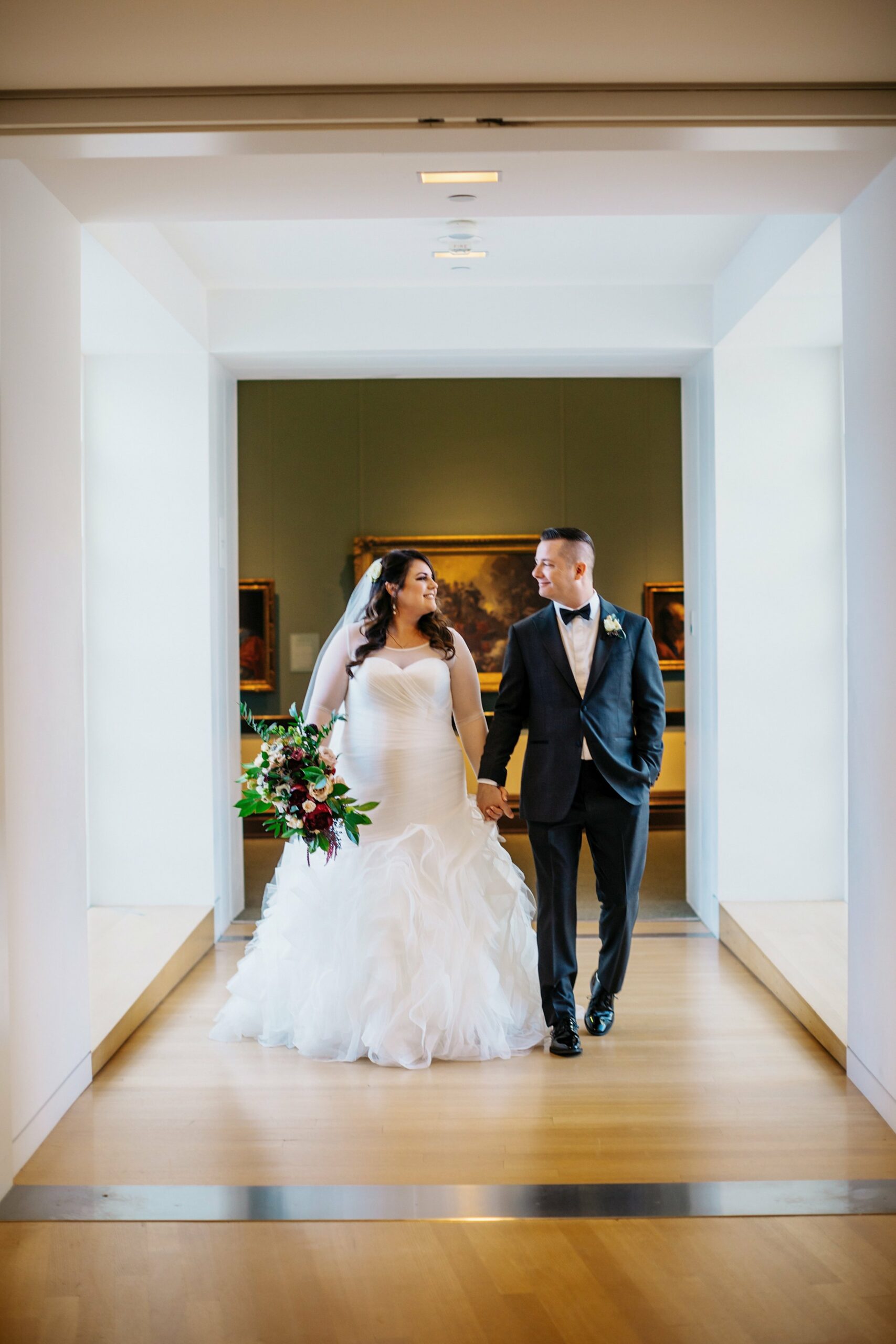 A bride in a white gown and groom in a tuxedo walk hand in hand down a brightly lit hallway. The bride holds a bouquet of red and white flowers.