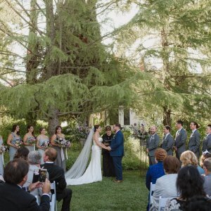 A bride and groom stand at the altar in an outdoor wedding ceremony, surrounded by their wedding party and attendees seated in white chairs.