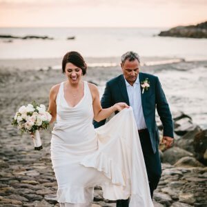 A couple, dressed in wedding attire, walks along a rocky beach at sunset. The bride holds a bouquet and lifts her dress slightly; the groom walks beside her, holding her train.