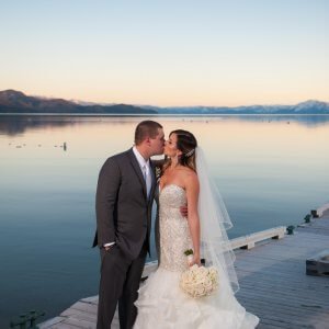 A bride and groom dressed in wedding attire share a kiss on a wooden dock by a lake with mountains in the background at sunset.