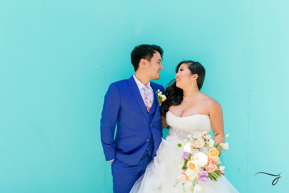 A bride and groom stand against a bright turquoise wall. The groom wears a blue suit, and the bride is in a white gown holding a bouquet of flowers. Both are looking at each other and smiling.