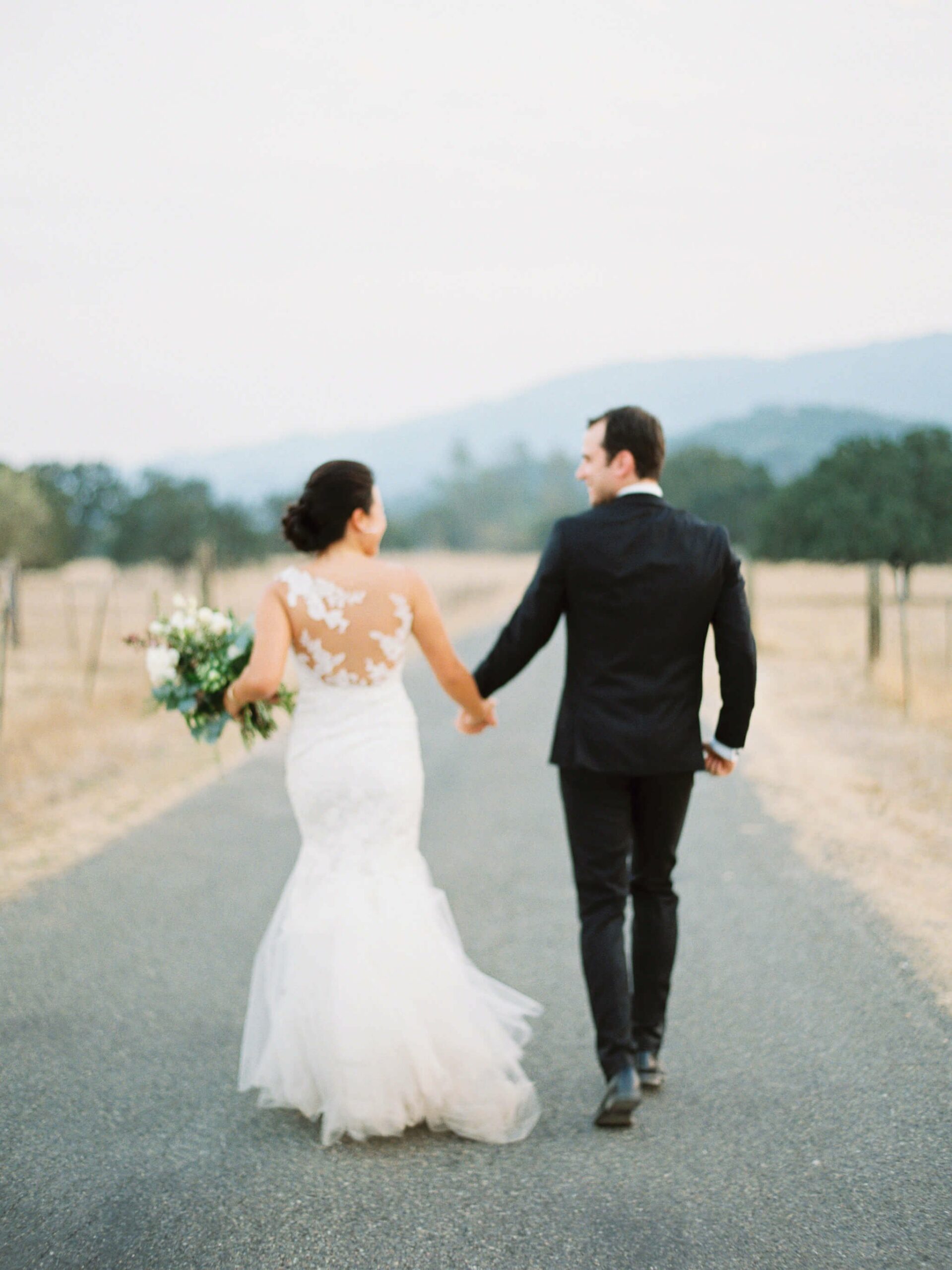 A couple in wedding attire walks hand in hand down a paved road. The bride holds a bouquet of flowers. The background features a landscape with distant hills and trees.