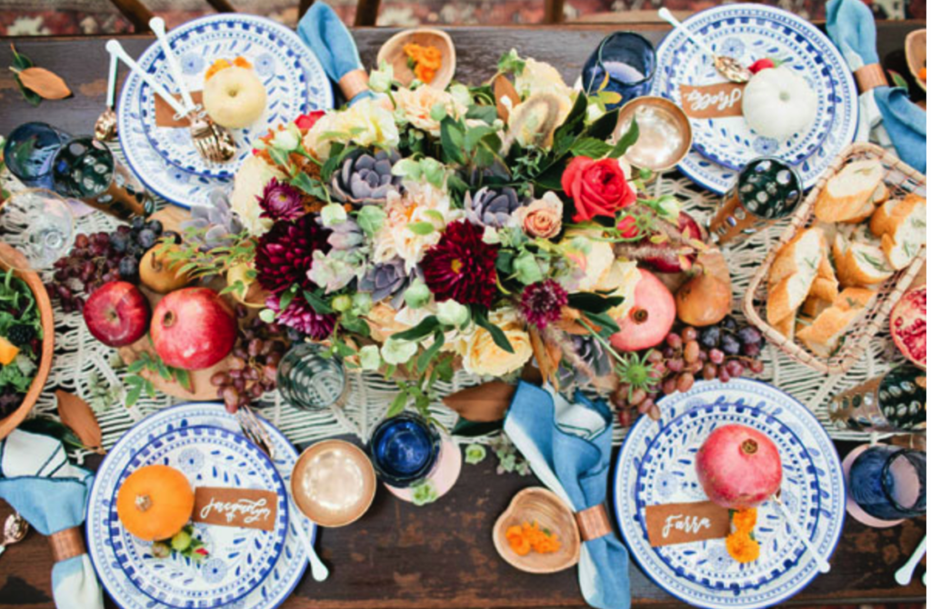 A table is set with colorful plates, cutlery, bread, fruit, and a central floral arrangement. The plates have name tags and decorations throughout give a festive and abundant feel.