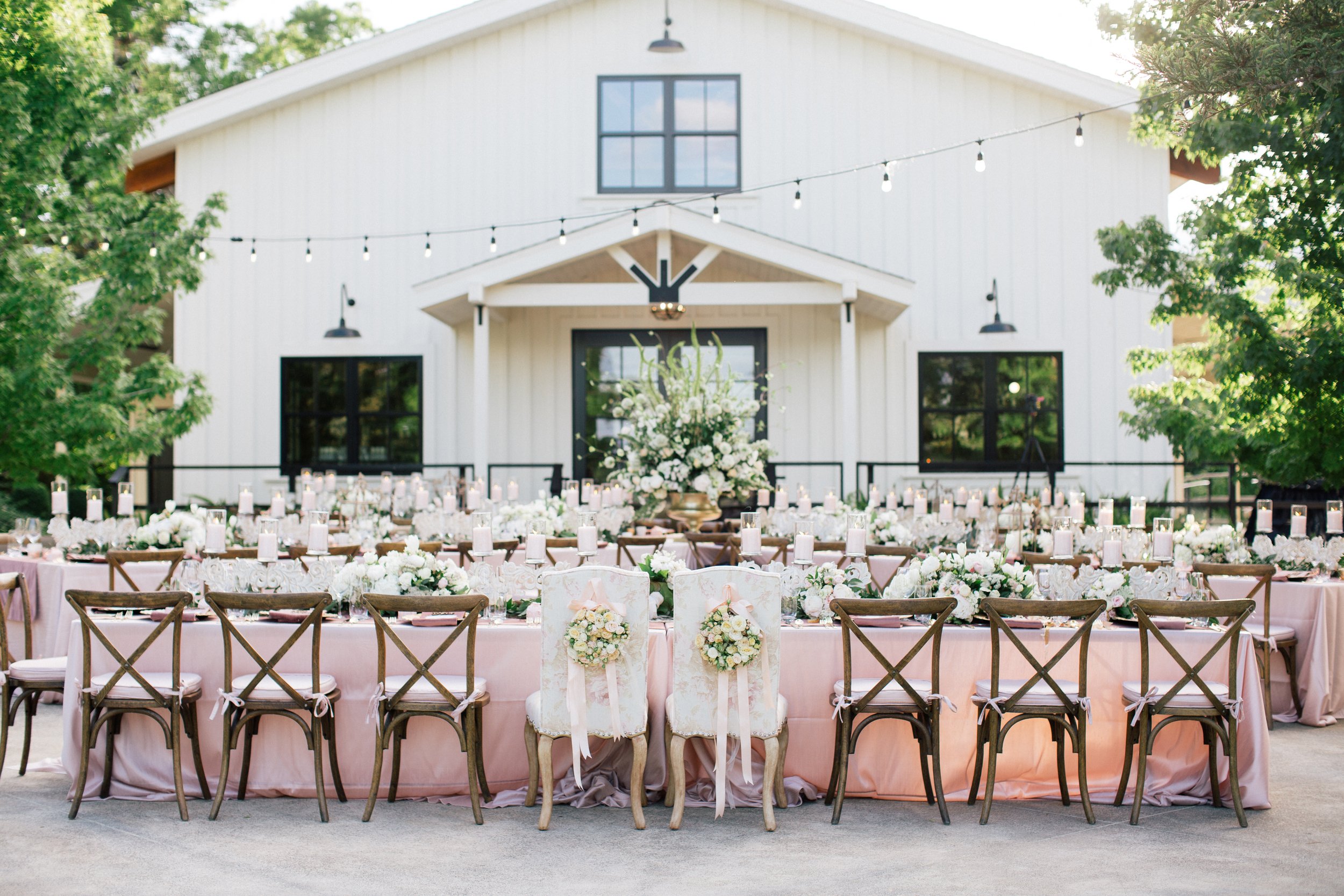 An outdoor wedding reception setup with long tables adorned with floral arrangements, candles, and greenery. A barn-like building is in the background, and string lights are hung above.